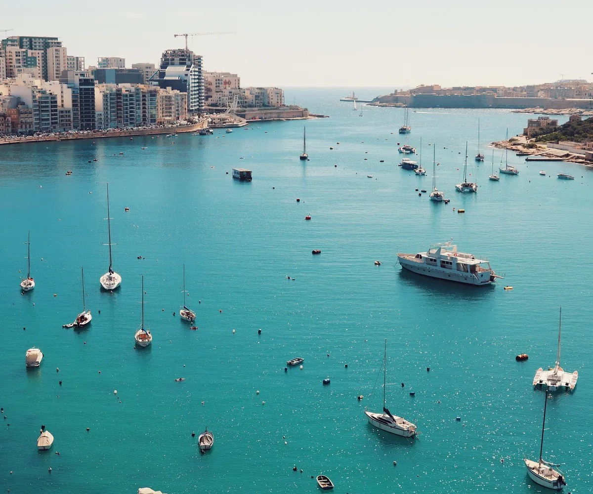 Sailing boats moored across a turquoise Maltese bay, with a modern apartment waterfront on one shore and a fortified headland on the other