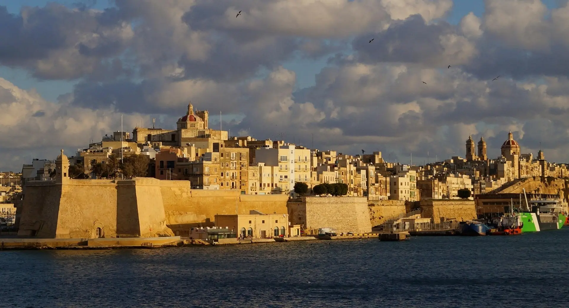Malta's fortified harbour at golden hour, honey-coloured limestone bastions and tiered townhouses rising above the water beneath a clouded sky